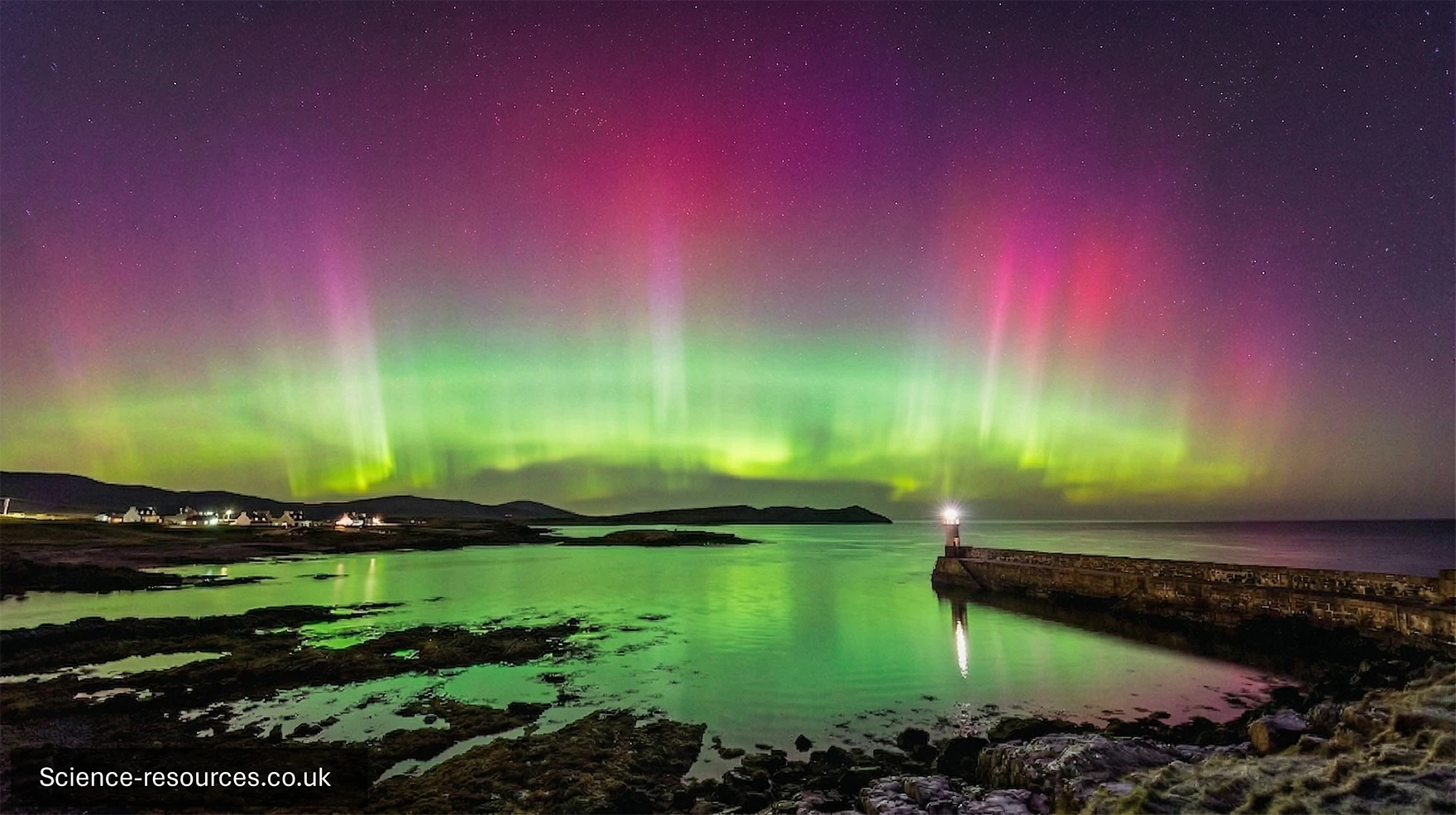 A vivid photograph captures the Northern Lights glowing over a quiet coastal landscape at night. The sky is filled with sweeping bands of green, pink, and deep red aurora light stretching across the horizon. The colours blend and rise in vertical curtains, creating a dramatic display against a dark, star‑speckled sky.
Below the aurora, calm water reflects the green glow, giving the sea a luminous sheen. A stone pier extends from the right side of the image, ending with a small beacon or lamp casting a soft white light onto the water. Rocky shoreline and pools of shallow water fill the foreground, while distant hills and a small cluster of lights from buildings sit on the left side, indicating a quiet coastal community. The overall scene is serene, colourful, and atmospheric, showing the aurora sweeping over a peaceful harbour at night.