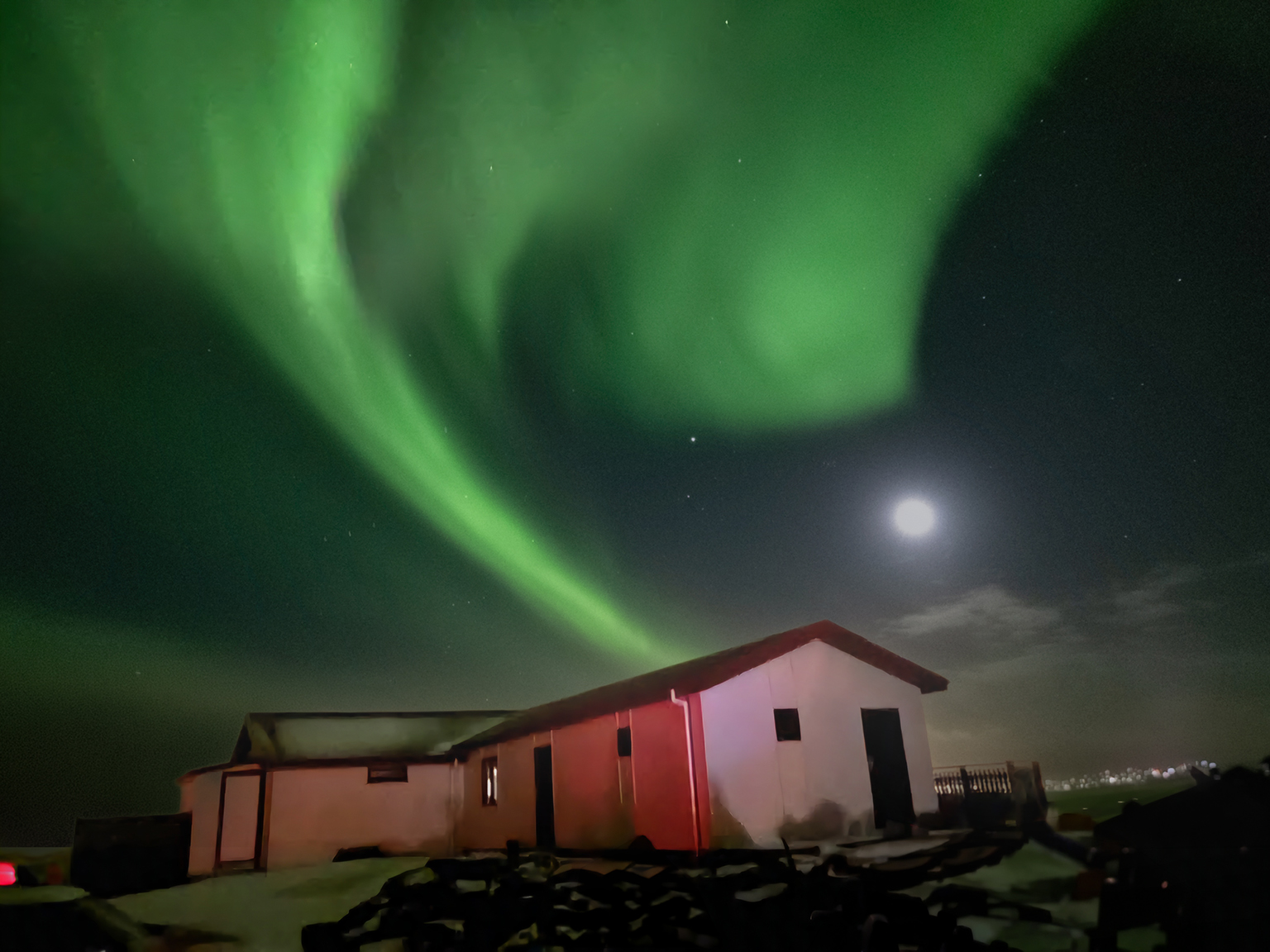 A night-time photograph taken in Iceland in March 2025, showing vivid green aurora borealis sweeping across the sky in wide, flowing arcs. The aurora forms a twisting, luminous ribbon that curves above a small, single‑storey house with a red roof and pale exterior walls. The house sits in a dark, rocky landscape illuminated faintly by the glow of the aurora and a bright full moon on the right. Stars are visible through gaps in the auroral light, and thin clouds hover near the horizon. The scene feels remote and peaceful, dominated by the striking natural light display overhead.